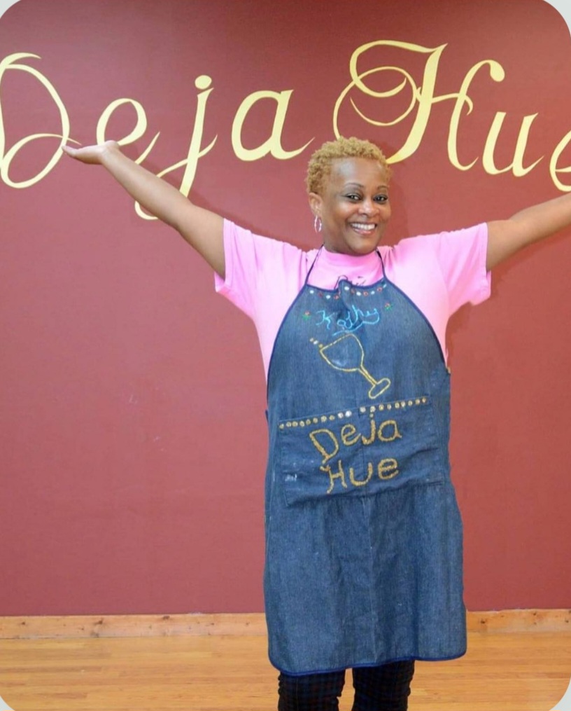 Kathryn Jackson in a Deja Hue Art apron standing in front of the Deja Hue Art studio wall